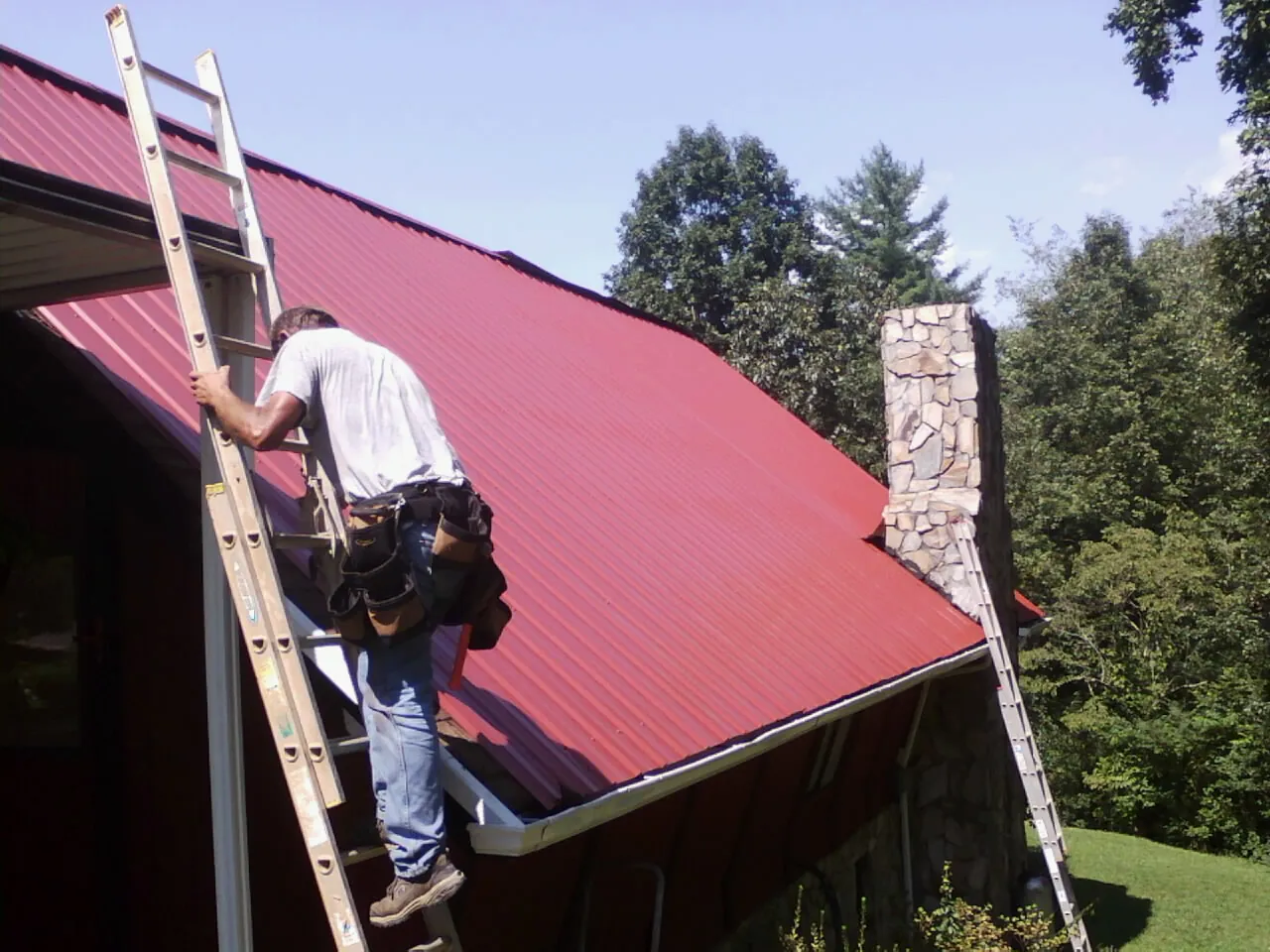 Roofer on a ladder beside a red metal roof and stone chimney — Soffit & Fascia Repair by Roofing Brothers in Youngstown
